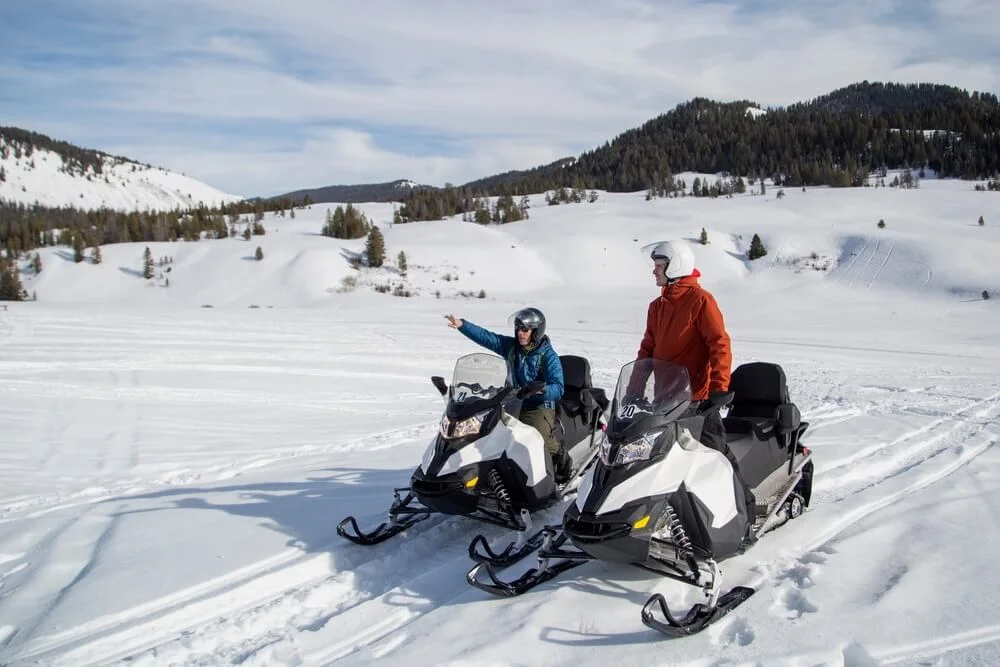 Two snowmobilers in helmets pausing on a snowy meadow below forested hills