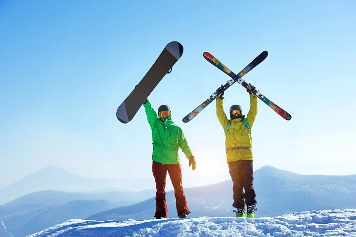 Skier and snowboarder holding their skis and snowboard overhead on a snowy mountain summit