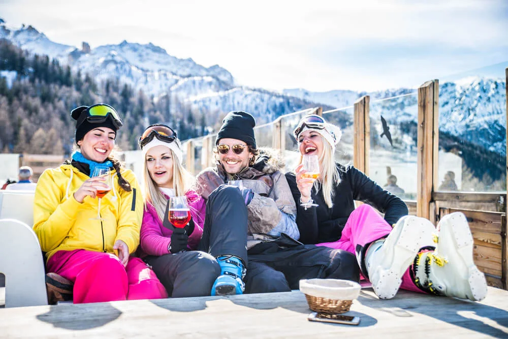 Friends in ski gear laughing and toasting with drinks on a sunny mountain deck